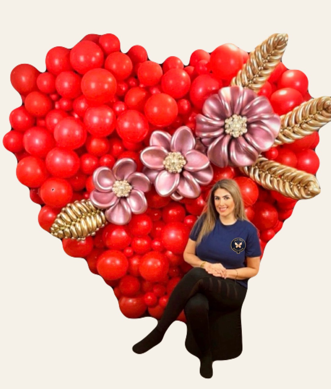 Grand cœur en ballons rouges orné de fleurs roses et de feuilles dorées, avec une femme assise devant